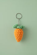 A crocheted carrot keychain with a green top and orange body, hanging from a metal ring on a textured off-white wall.