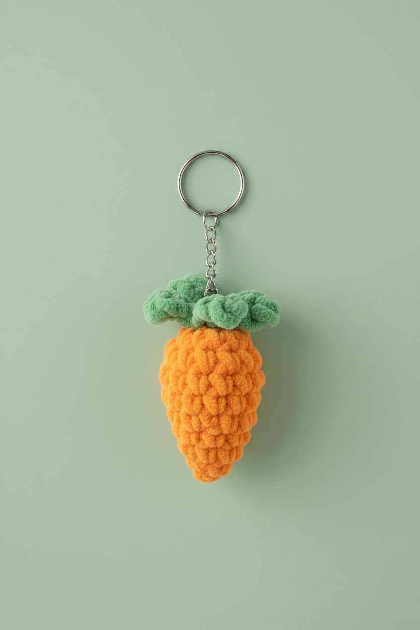 A crocheted carrot keychain with a green top and orange body, hanging from a metal ring on a textured off-white wall.