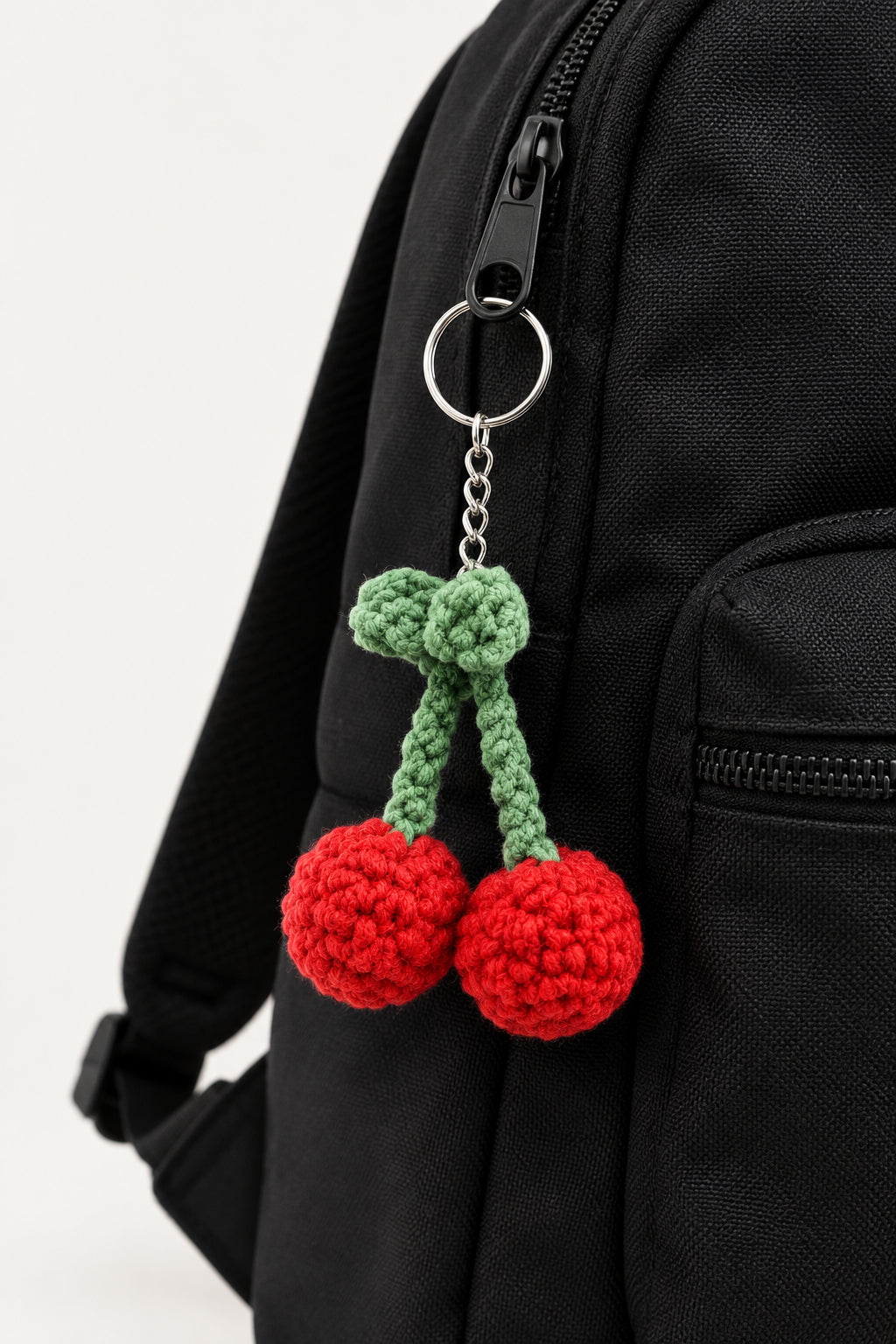 A crocheted cherry keychain featuring two red yarn pom-pom cherries with green stems and a silver metal key ring, shown against a textured off-white wall.