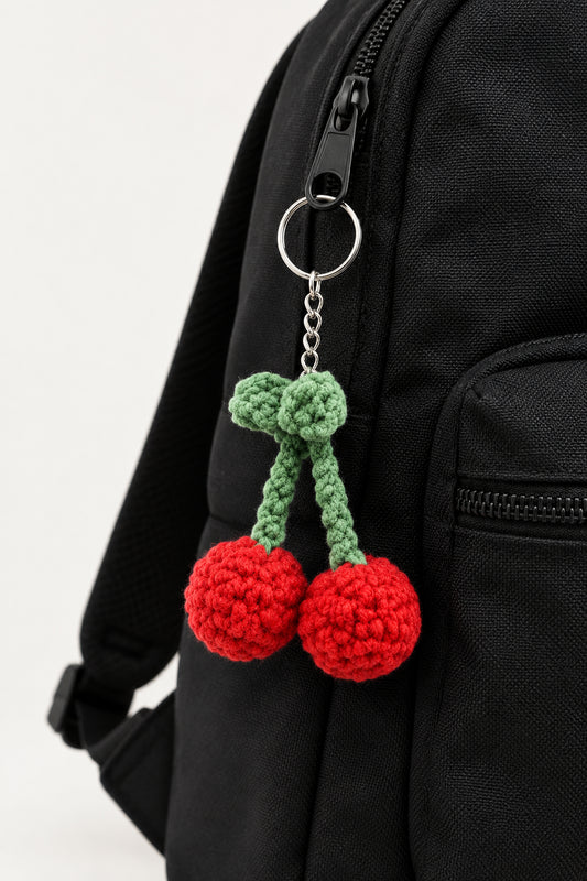 A crocheted cherry keychain featuring two red yarn pom-pom cherries with green stems and a silver metal key ring, shown against a textured off-white wall.