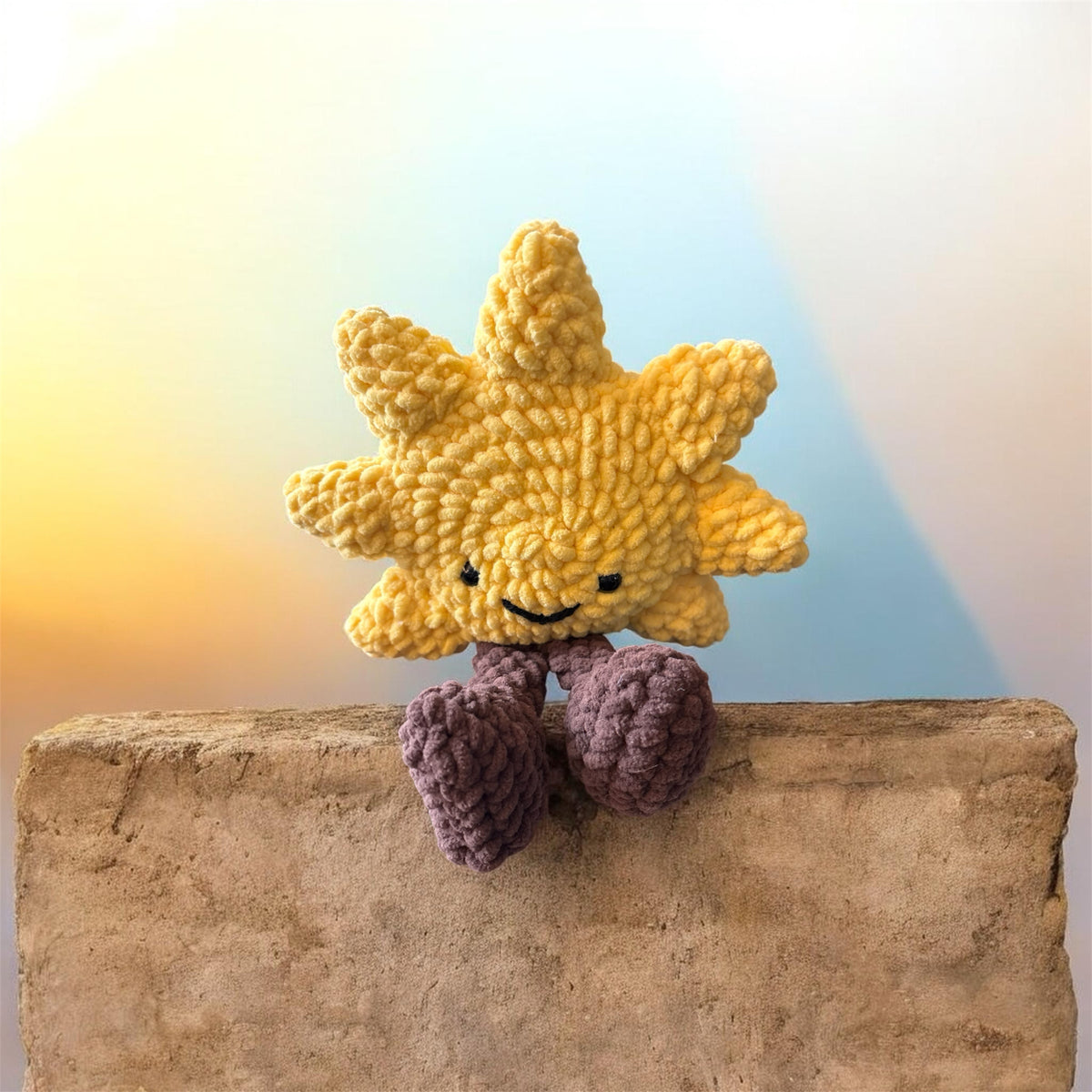 Hook Happy Co. handmade yellow crochet sun plush toy with a smiling face and dangling brown legs sitting on a ledge.